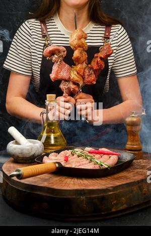 Frau mit unterschiedlichem Fleisch auf Spieß steht vor einer Pfanne mit Würstchen Stockfoto