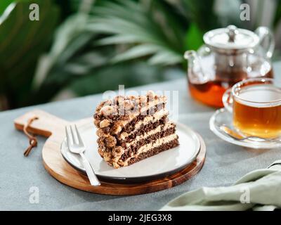 Perfekter ästhetischer Schichtenkuchen mit Karamell und Nüssen auf dem Teller über dem grauen Tisch. Stück Schokoladenmandelkeks mit Karamell-Zuckerguss in der Nähe von Teetasse und Teekannen. Stück Biskuitkuchen im Café Stockfoto