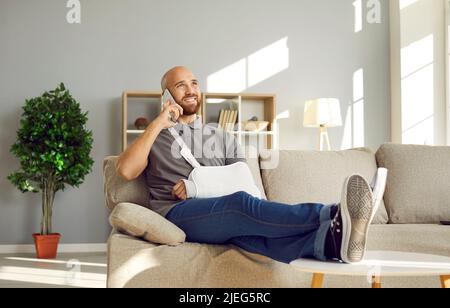 Entspannter Mann mit gebrochenem Arm, der Spaß hat, während der Rehabilitation zu Hause mit Freunden zu telefonieren. Stockfoto