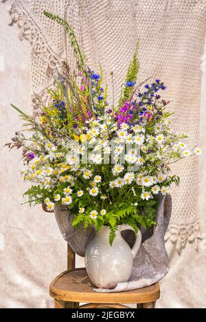 Strauß Wildblumen mit Kamillen, Kornblumen, Fuchshandschuh, ivan-Tee, Kräuter in einem weißen Krug auf einem Stuhl. Arrangement von einem Floristen gemacht. Stockfoto