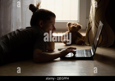 Das Mädchen liegt auf dem Boden und arbeitet auf einem Laptop in einem dunklen Raum neben dem Fenster, arbeitet und chatet online zu Hause Stockfoto