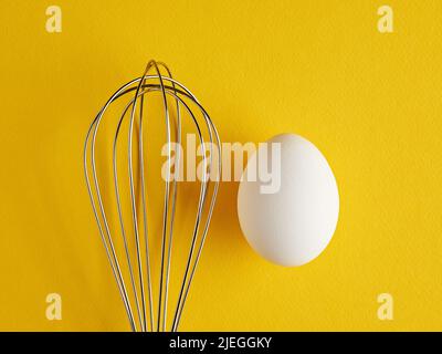 Ein Whisker und ein weißes Ei auf gelbem Hintergrund. Rührbesen zum Kochen von Eierquirlen. Stockfoto