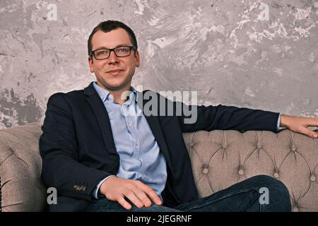 Porträt eines jungen kaukasischen lächelnden Geschäftsmannes in Jacke und Brille, der in offener Position auf dem Sofa sitzt und gegen die Kamera blickt Stockfoto