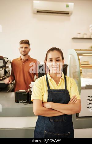 Lächelnder, selbstbewusster Kellner im Coffeeshop, der mit gekreuzten Armen steht, Barista, der im Hintergrund arbeitet Stockfoto