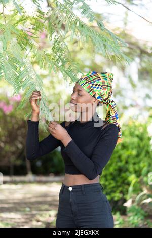 Schönes afrikanisches Mädchen in schwarz gekleidet mit einem farbigen Kopftuch, das im Garten steht und aufmerksam auf die Blätter eines Mimosenbaums schaut; Naturliebhaber Stockfoto