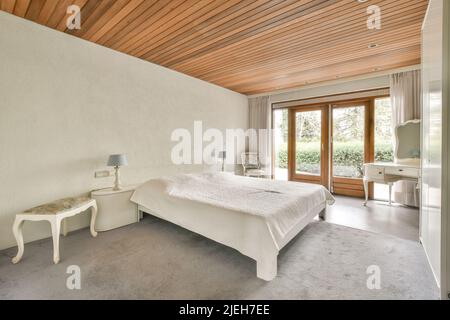 Komfortables Bett mit warmer Decke in der Nähe von Fenster und Tür, die zur Terrasse führt, in einem stilvollen Schlafzimmer mit grauen Wänden Stockfoto