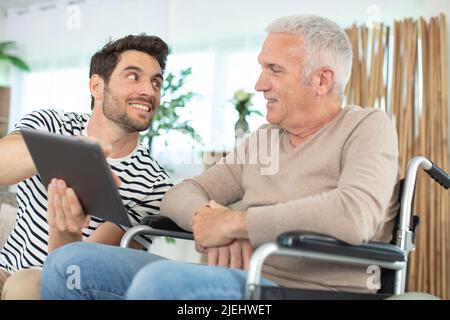 Fröhlicher behinderter Rentner, der im Rollstuhl in der Nähe des bärtigen Sohnes sitzt Stockfoto