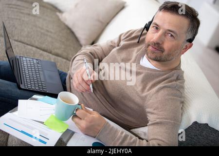 Mann, der auf der Couch sitzt und mit einem Laptop und einem Headset studiert Stockfoto