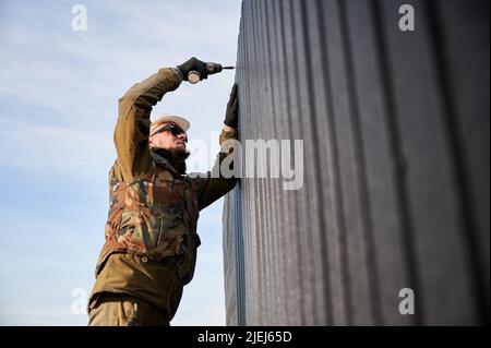 Männlicher Baumeister, der schwarze Wellblech installiert, das als Fassade des zukünftigen Häuschens verwendet wird. Mann Arbeiter Gebäude Holzrahmen Haus. Zimmerei- und Konstruktionskonzept. Stockfoto