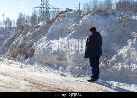 Ein Mann, der neben einem großen Schneehaufen auf der Straße steht. Hohe Schneeverwehungen nach Schneefall oder Schneesturm. Schneeräumung auf Straßen für sicheres dri Stockfoto