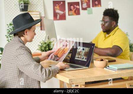 Junge professionelle Redakteurin untersucht Fotomodell sitzen an ihrem Arbeitsplatz, während retuschieren Fotos auf Laptop Stockfoto