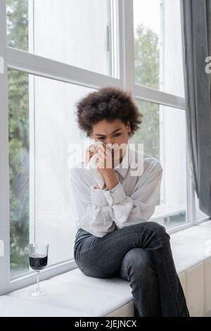 Traurige afroamerikanische Frau, die Tränen in der Nähe eines Glases Rotwein wischt, während sie auf dem Fensterbrett sitzt Stockfoto
