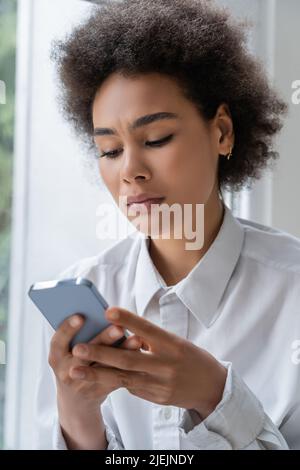 Traurige afroamerikanische Frau im weißen Hemd, die Nachricht auf dem Smartphone liest Stockfoto