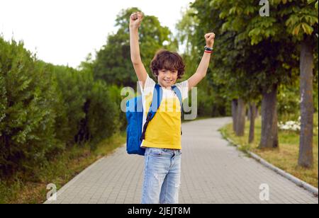 Fröhlicher, fröhlicher kleiner Schuljunge, der im Park steht, seine Hände hochhebt und lächelt Stockfoto