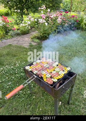 Fleischsteaks mit Gemüsestückchen werden auf einem Gitter auf dem Grill im Garten vor dem Hintergrund von Rasen, Rosen und einem Gartenweg gebraten. Stockfoto