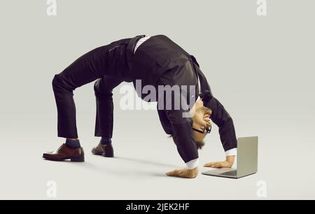 Glücklicher flexibler Geschäftsmann im Anzug, der Yoga macht, während er an seinem Laptop arbeitet Stockfoto