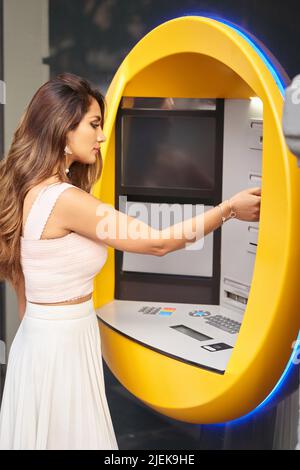 Eine Frau führt eine Kreditkarte in einen Geldautomaten ein, während sie draußen auf der Straße steht. Geld-, Banken- und Urban-Lifestyle-Konzept. Stockfoto