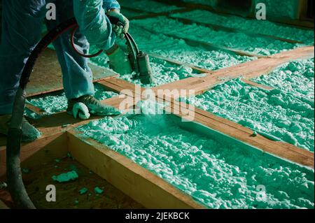 Männlich Baumeister isolierenden Holzrahmen Haus. Beschnittene Ansicht eines Arbeiters, der Polyurethan-Schaum auf den Boden in der zukünftigen Hütte sprüht, mit Plural Component Gun. Bau- und Isolierkonzept. Stockfoto