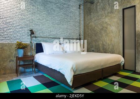 Schlafzimmer mit Betonwand und Ziegelwand, Teppich, Bett, Lampe und Vase mit getrockneten Blumen Stockfoto