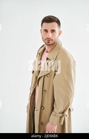 Porträt eines jungen trendigen Mannes in beigem Trenchcoat auf weißem Studiohintergrund Stockfoto