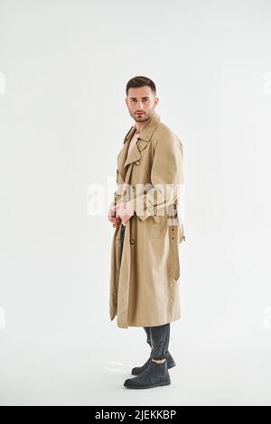 Porträt eines jungen trendigen Mannes in beigem Trenchcoat auf weißem Studiohintergrund Stockfoto