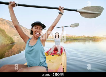 Portrait zwei junge Frauen jubeln und feiern beim Kanufahren auf einem See. Begeisterte Freunde, die unterwegs Rudern und Kajakfahren auf einem Fluss genießen Stockfoto