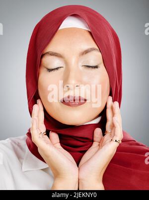 Eine schöne junge muslimische Frau mit geschlossenen Augen, die rotes Kopftuch und Lippenstift vor grauem Studiohintergrund trägt. Bescheidene arabische Frau, die Make-up trägt Stockfoto