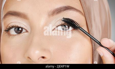 Nahaufnahme Porträt einer muslimischen Frau, die Mascara-Make-up auf die Wimpern aufgetragen hat. Kopfschuss eines atemberaubenden arabischen Models, das ihre Schönheitsroutine gegen Grau macht Stockfoto