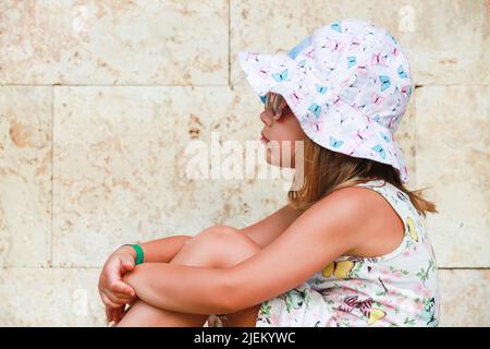 Nette europäische Baby-Mädchen in bunten Panama-Hut und Sonnenbrille sittingg in der Nähe der Steinwand, Profil Porträt Stockfoto