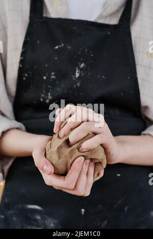 Hände junger weiblicher Kunsthandwerkerin in schwarzer Schürze, die ein Stück Ton kneten, um Steingut oder andere kreative Tonwaren zum Verkauf zu machen Stockfoto