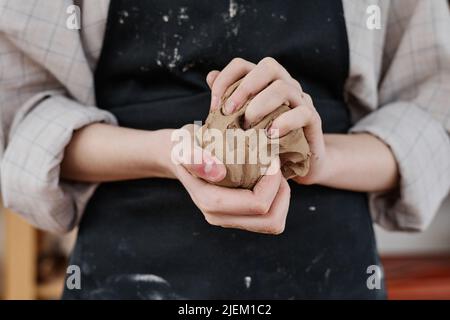 Hände einer jungen kreativen Frau in Arbeitskleidung, die ein Stück weichen Tones hält und knetet, während sie sich für die Herstellung von Steingut-Produkten vorbereitet Stockfoto