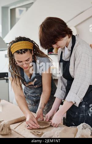 Junge weibliche Tutorin oder Meister biegen über Tisch mit abgeflachten Stück Ton helfen Mädchen in Schürze Steingut Artikel in der Werkstatt zu schaffen Stockfoto