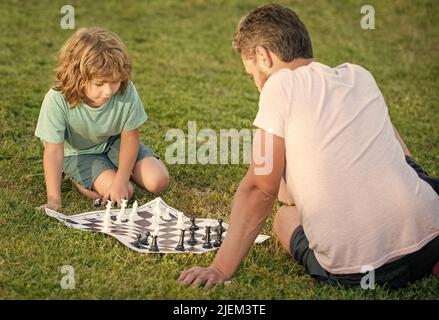 Glückliche Familie von Vater Mann und Sohn Junge spielen Schach auf grünem Gras im Park im Freien, Gegner Stockfoto