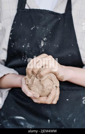 Hände einer jungen kreativen Frau in Schürze kneten Stück weichen Ton, bevor neue handgemachte Steingut-Gegenstände in der Werkstatt zu schaffen Stockfoto