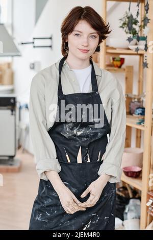 Junge kreative Kanne in Arbeitskleidung, die vor der Kamera gegen das Innere der Werkstatt oder des Studios und Holzregale steht Stockfoto