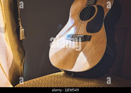 Eine spanische akustische klassische Gitarre mit Stahl- und Nylonsaiten, die auf einem Stuhl neben einem Fenstervorhang ruht Stockfoto