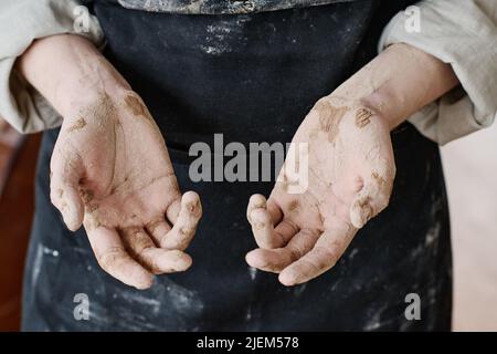 Offene Handflächen mit trockenem Lehm von junger, erfahrener Kunsthandwerkerin in schwarzer Schürze, die vor der Kamera steht und ihre Hände zeigt Stockfoto