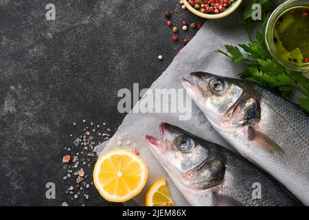 Seebarsch roh. Frischer Meeresfischbarsch mit Salz, Pfeffer, Petersilie, Olivenöl und Zitrone auf Pergamentpapier auf dunklem Beton-rustikalem Hintergrund. Essen kochen Stockfoto
