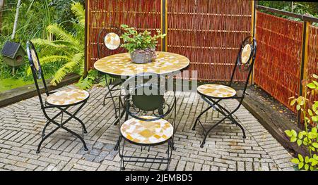 Set von Essmöbeln im Freien. Mosaikfliesen und eiserne Stühle und Tisch auf einer grauen Backsteinterrasse in einem üppigen Garten. Gemütlicher geschlossener Entspannungsbereich draußen Stockfoto