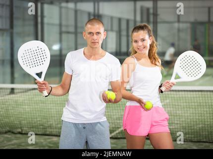 Ein paar fröhliche junge Paddle-Tennisspieler stehen auf dem Hallenplatz Stockfoto