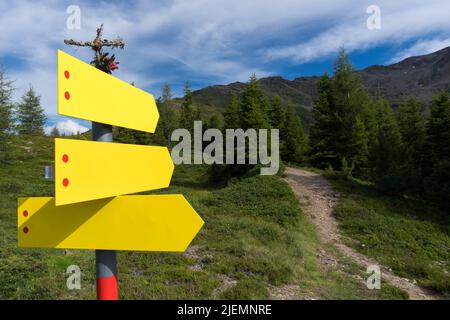 Drei deutlich sichtbare gelbe Wegweiser zeigen in verschiedene Richtungen neben einem schmalen Fußweg in alpinem Gelände unter teilweise bewölktem, blauem Himmel Stockfoto