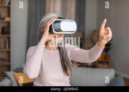 Fröhliche europäische ältere Frau mit grauen Haaren in vr Brille spielt in Online-Spiel zur freien Zeit im Zimmer Innenraum Stockfoto