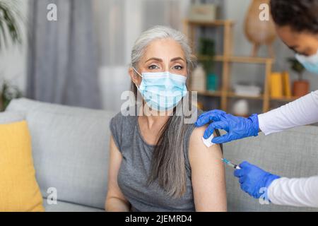 Arzt in Schutzhandschuhen macht Injektion der europäischen alten Frau in Maske mit grauen Haaren im Wohnzimmer Stockfoto