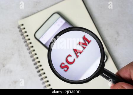 Die Hand eines Mannes hält eine schwarze Lupe am Griff auf einem Smartphone-Hintergrund mit Betrug Text. Finanz- und Geschäftskonzept. Stockfoto