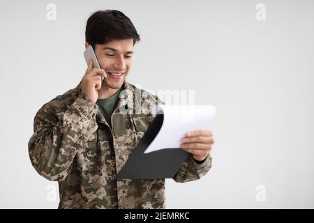 Lächelnder junger Militärangehöriger hält Chart und telefoniert Stockfoto