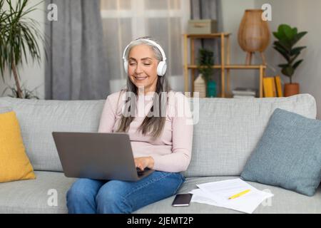 Die glückliche europäische alte Frau mit grauen Haaren in Kopfhörern hat sich aus der Ferne treffen lassen, hört Musik und schaut sich Videos an Stockfoto