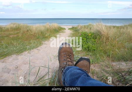 Mann mit ausgestreckten Beinen auf Sanddüne, Paar Füße und Jeans auf einem Hintergrund aus grünem Gras und blauer Meeresoberfläche Stockfoto