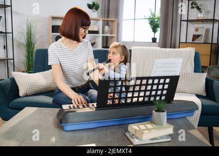 Kleines nettes Mädchen mit Musiklehrerin, die an der Musikschule Flötenunterricht hat. Lächelnde junge Frau mit rotem Haar, die auf ein kleines Mädchen schaut, das auf dem Sofa sitzt und im Klassenzimmer Klavier spielt. Stockfoto