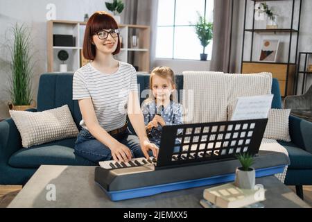 Kleines nettes Mädchen mit Musiklehrerin, die an der Musikschule Flötenunterricht hat. Lächelnde junge Frau mit rotem Haar, die auf ein kleines Mädchen schaut, das auf dem Sofa sitzt und im Klassenzimmer Klavier spielt. Stockfoto