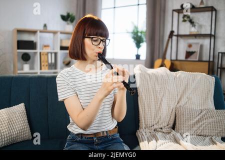 Die junge Frau spielt zu Hause im hellen Wohnzimmer Flöte. Mädchen sitzt auf dem Sofa mit einem Musikinstrument drinnen. Hübsche junge Frau studiert Flöte spielen. Stockfoto